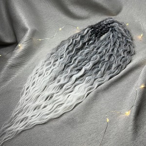 Op de afbeelding: Een lange, golvende haarverlenging met een ombre kleurovergang van zwart naar wit. Het haar is gestyled in losse golven en wordt getoond tegen een grijze stoffen achtergrond.