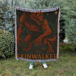 May include: A woven throw blanket featuring a red werewolf illustration against a dark green background with trees and stars. The word "SKINWALKER" is printed in a brown font at the bottom. The blanket has fringed edges.