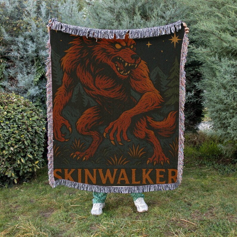 May include: A woven throw blanket featuring a red werewolf illustration against a dark green background with trees and stars. The word "SKINWALKER" is printed in a brown font at the bottom. The blanket has fringed edges.