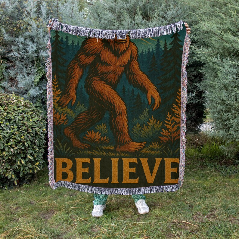 Op de afbeelding: Een geweven deken met een bruine Bigfoot die door een bos loopt. De deken heeft een donkergroene achtergrond met het woord "BELIEVE" in gouden letters. De randen zijn gefranjerd.