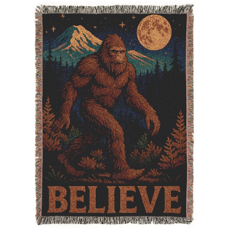 Op de afbeelding: Geweven sprei met een bruine Bigfoot-figuur die in een bos loopt onder een volle maan en een sterrenhemel. Het woord "BELIEVE" staat onderaan. De deken heeft een gefranjerde rand. Donker kleurenpalet.