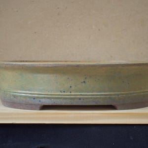 以下が含まれることがあります： 手作りの楕円形の盆栽鉢。セラミック製の鉢は、明るい緑色と茶色の釉薬に濃い青色の斑点があります。鉢の長さは約30cm、幅は約20cmで、ロープロファイルで長方形の足が付いています。