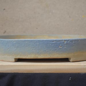 May include: An oval-shaped ceramic bonsai pot featuring a textured, matte light blue glaze on the upper portion and a natural, unglazed beige clay finish on the lower section, supported by small, integrated feet along the base.