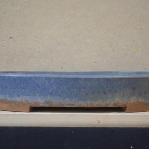 Op de afbeelding: Een ondiepe, ovaalvormige keramische bonsaischaal met een blauw geglazuurde binnen- en buitenkant, die overgaat in een ongeglazuurde, getextureerde terracotta-kleurige basis met kleine voetjes voor verhoging en drainage.