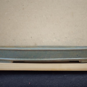 Pode incluir: Um vaso de bonsai oval feito à mão. O vaso de cerâmica tem um esmalte verde-azulado suave com uma borda mais escura. O vaso tem aproximadamente 30 cm de comprimento e está sobre uma prateleira de madeira contra um fundo neutro.