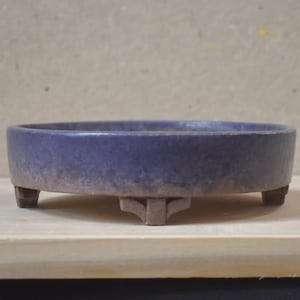 Peut inclure: Un pot à bonsaï rond et peu profond en céramique, doté d'une finition émaillée bleu foncé sur la partie supérieure et d'une base en argile brune naturelle non émaillée. Le récipient repose sur trois petits pieds robustes.