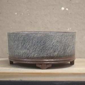 May include: A round, low-profile ceramic bonsai pot featuring a textured, mottled grey exterior surface with a dark brown, smooth-finished base rim. The vessel is supported by four small, evenly spaced feet and has a wide, open top opening.