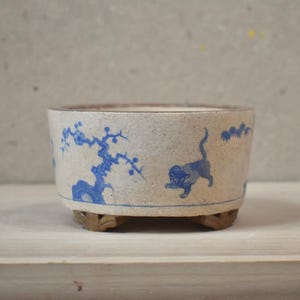 May include: A round, off-white ceramic bonsai pot featuring blue painted illustrations of a small cat and a flowering tree. The pot has a textured, matte finish and is supported by three small, brown, carved wooden feet at the base.