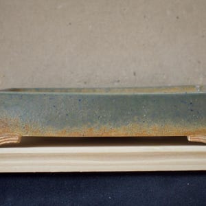 May include: A rectangular bonsai pot with a blue-green glaze and a rustic, weathered appearance. The pot has a light-coloured wooden base and is designed for planting small trees or shrubs. The pot is approximately 30 cm long.