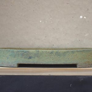 May include: A shallow, oval-shaped ceramic bonsai pot with a textured, mottled surface in shades of muted green and earthy brown. The container features a low profile with a flat rim and a small, rectangular cutout at the base for drainage.