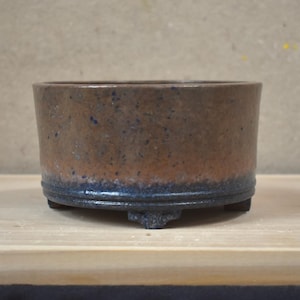 May include: A cylindrical ceramic pot with a flat rim and three small feet. The exterior features a mottled brown and tan upper section that transitions into a dark, deep blue glazed base, creating a distinct two-tone color gradient effect.