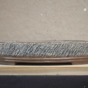 May include: A shallow, oval-shaped ceramic bonsai pot featuring a textured exterior with a grey and black cross-hatched pattern. The base of the pot is a smooth, solid brown clay with a raised rim and small feet for elevation.