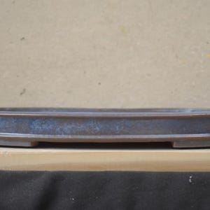May include: An oval ceramic bonsai pot with a low profile and a raised rim. The exterior features a dark brown base color with a mottled, dusty blue glaze finish. The pot has a flat bottom with small feet and a smooth, matte-like texture.