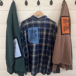 May include: Three plaid shirts hanging on hooks. The center shirt is blue and black with a blue patch on the back. The patch has a black and white design of a face with mushrooms and leaves. The shirt on the left is green with a white and blue patch on the sleeve. The shirt on the right is brown and white with an orange patch on the sleeve. The patch has a black and white design of a face with mushrooms and leaves.