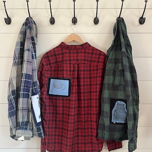 May include: Three plaid shirts hanging on hooks on a white wall. The center shirt is red and black with a blue and white patch on the left sleeve. The shirt on the left is blue and white plaid. The shirt on the right is green and black plaid with a blue and white patch on the right sleeve.
