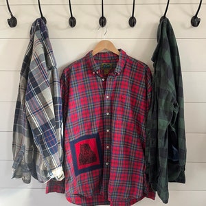 May include: A red and black plaid flannel shirt with a bear patch on the pocket. The shirt is hanging on a hook with two other plaid shirts.