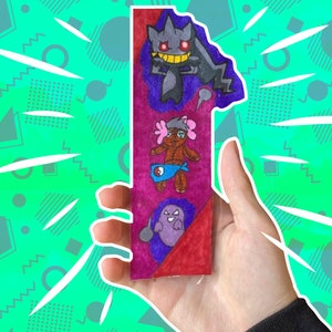 May include: A hand holds a bookmark with a red, purple and blue background. The bookmark features three cartoon characters: a purple ghost with a red eye, a brown teddy bear with a pink bow, and a purple and black ghost with red eyes.