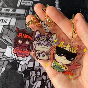 May include: Three clear acrylic keychains with cartoon character designs. The keychains have gold-coloured hardware. The characters are in red, purple, and orange with text that reads "AH AH", "OCURE", and "ROYALLY PUMPY".