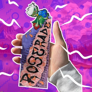 May include: A hand holds a bookmark with a colorful, abstract design and the word "ROGUEGRAD" written in a stylized font. A cartoon character with green and blue accents is perched on top of the bookmark.