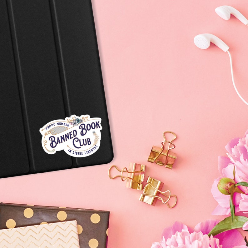May include: A white sticker with a purple and gold design that says "Proud Member Banned Book Club In Libris Libertas".
