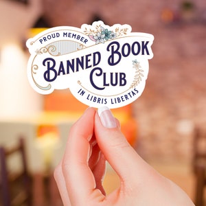 May include: A white sticker with a gold border and floral accents. The sticker says "Proud Member Banned Book Club In Libris Libertas".