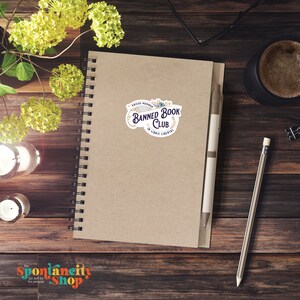 May include: A brown notebook with a white sticker that says "Proud Member Banned Book Club In Libris Libertas" with floral accents. The sticker is on a spiral bound notebook.