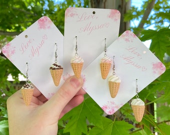 May include: Three pairs of ice cream cone earrings on white cards with pink floral designs. The cards have the text "Love, Alyssa" on them. The earrings are silver with a white and brown ice cream cone charm.