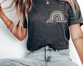 Puede incluir: Una camiseta de color gris jaspeado oscuro con un arcoíris y el texto "Mrs. Johnson" impreso en la parte delantera.