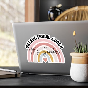 May include: A rainbow-colored sticker with the text "Instructional Coach Ms. Sanders" and a small heart in the center. The sticker is on a silver laptop.