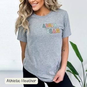 Op de afbeelding: Een grijs T-shirt met de tekst "ADMIN Team" in kleurrijke, retro-stijl letters. De tekst staat op de voorkant van het shirt, gecentreerd op de borst.