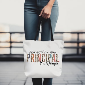 Op de afbeelding: Witte tote bag met zwarte handvatten. De tas heeft de tekst "Alphabet Elementary PRINCIPAL Ms. Stringer" erop gedrukt. Het woord "PRINCIPAL" staat in kleurrijke letters met een luipaardprint.