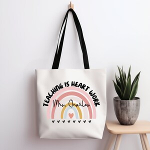 Może przedstawiać: Biała torba typu tote z czarnymi uchwytami, przedstawiająca tęczę i napis "Teaching is heart work" z imieniem "Mrs. Charles" w środku tęczy.