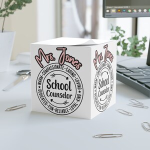 Puede incluir: Caja blanca en forma de cubo con un diseño rosa y negro. La caja tiene el texto "Mrs. Jones" y "School Counselor". El texto está rodeado por un círculo con las palabras "Warm-Compassionate-Caring-Loving-Dedicated-Fun-Reliable-Loyal-Kind" alrededor.