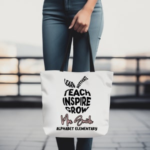 Op de afbeelding: Witte tote bag met zwarte handvatten en een zwart appelontwerp. De appel is gemaakt van de woorden "Leren, Motiveren, Leren, Inspireren, Groeien". Onder de appel staat de tekst "Mrs. Smith Alphabet Elementary" in roze en zwart.