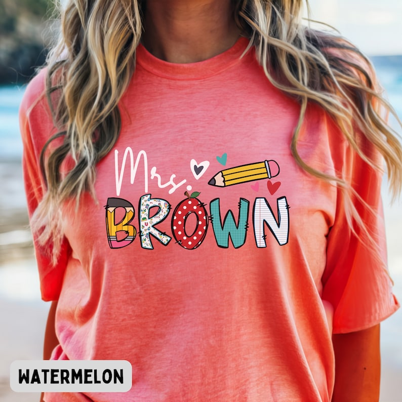 Może przedstawiać: Koralowa koszulka z napisem "Mrs. BROWN" w zabawnym, kolorowym wzorze. Projekt zawiera oł&oacute;wek, serca i jabłko. Słowo "WATERMELON" jest wydrukowane na dole koszulki.