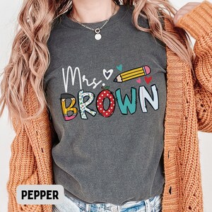 Może przedstawiać: Ciemnoszara koszulka z napisem "Mrs. Brown" w kolorowym, zabawnym wzorze. Wz&oacute;r zawiera oł&oacute;wek, serca i jabłko. Koszulka jest w wygodnym, swobodnym stylu.