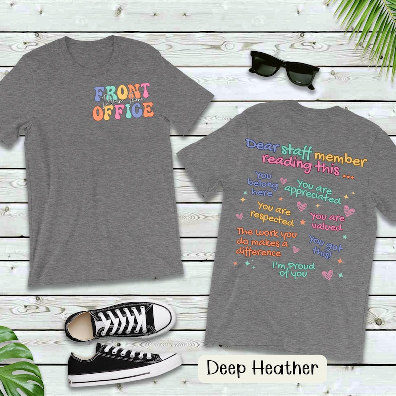 K&ouml;nnte beinhalten: Dunkelgraue T-Shirts mit dem Text "FRONT OFFICE" in bunten Buchstaben auf der Vorderseite. Auf der R&uuml;ckseite des Shirts steht der Text "Liebe/r Mitarbeiter/in, der/die dies liest..." mit positiven Affirmationen.
