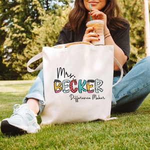 Może przedstawiać: Biała torba z płótna z napisem "Mrs. Becker Difference Maker" w kolorowych literach. Litery są ozdobione ołówkiem, jabłkiem i innymi wzorami w szkolnym stylu.