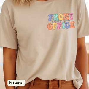 K&ouml;nnte beinhalten: Beiges T-Shirt mit dem Schriftzug "FRONT Dream Team OFFICE" in bunten Retro-Lettern auf der linken Brust. Das Shirt hat kurze &Auml;rmel und eine lockere Passform. Das Wort "Natural" ist auf einem wei&szlig;en Rechteck unten aufgedruckt.