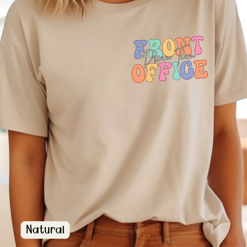 K&ouml;nnte beinhalten: Beiges T-Shirt mit dem Schriftzug "FRONT Dream Team OFFICE" in bunten Retro-Lettern auf der linken Brust. Das Shirt hat kurze &Auml;rmel und eine lockere Passform. Das Wort "Natural" ist auf einem wei&szlig;en Rechteck unten aufgedruckt.