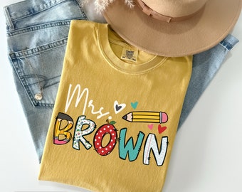 May include: Mustard-colored t-shirt with the text "Mrs. BROWN" in colorful, playful lettering, featuring hearts, a pencil, and an apple design. The shirt is folded and placed on top of blue jeans and a tan hat.