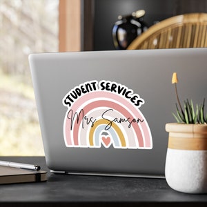 May include: A rainbow-colored sticker with the text "Student Services Mrs. Samson" and a small heart in the center. The sticker is on a silver laptop.