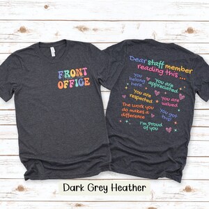 K&ouml;nnte beinhalten: Dunkelgraue Heather-T-Shirts mit farbenfrohem Text. Ein Shirt zeigt "FRONT OFFICE" in Retro-Schrift. Das andere Shirt hat den Text "Dear staff member reading this..." mit positiven Affirmationen. Die Shirts werden auf hellem Holzgrund gezeigt.