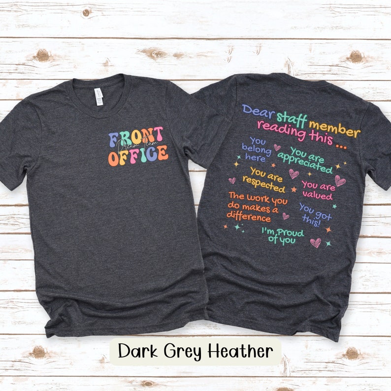 K&ouml;nnte beinhalten: Dunkelgraue Heather-T-Shirts mit farbenfrohem Text. Ein Shirt zeigt "FRONT OFFICE" in Retro-Schrift. Das andere Shirt hat den Text "Dear staff member reading this..." mit positiven Affirmationen. Die Shirts werden auf hellem Holzgrund gezeigt.