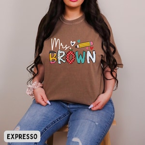 Może przedstawiać: Brązowa koszulka z napisem "Mrs. Brown" w kolorowych literach i grafiką oł&oacute;wka. Koszulka jest w jednolitym kolorze i ma luźny kr&oacute;j. Słowo "EXPRESSO" znajduje się na dole obrazu.