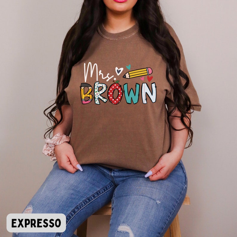 Może przedstawiać: Brązowa koszulka z napisem "Mrs. Brown" w kolorowych literach i grafiką oł&oacute;wka. Koszulka jest w jednolitym kolorze i ma luźny kr&oacute;j. Słowo "EXPRESSO" znajduje się na dole obrazu.