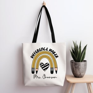 Op de afbeelding: Witte canvas tote bag met zwarte handvatten. De tas heeft een regenboog gemaakt van gele potloden met een zwart-wit hart in het midden. De tekst "Difference Maker" staat in het zwart boven de regenboog en "Mrs. Johnson" staat in het zwart onder de regenboog.
