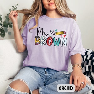 Może przedstawiać: Jasnofioletowa koszulka z napisem "Mrs. Brown" w kolorowym, zabawnym wzorze. Wz&oacute;r zawiera oł&oacute;wek, serca i słowo "BROWN" w r&oacute;żnych wzorach. Koszulka jest w wygodnym, swobodnym stylu.