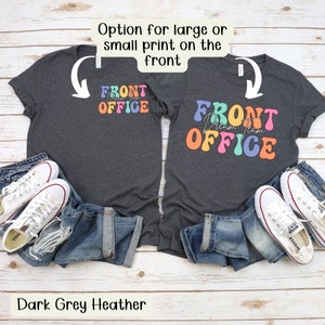 K&ouml;nnte beinhalten: Zwei dunkelgraue Heather-T-Shirts mit dem Text "FRONT OFFICE Dream Team OFFICE" in einer farbenfrohen Retro-Schriftart. Die Shirts sind mit blauen Jeans und wei&szlig;en Turnschuhen kombiniert. Das Bild enth&auml;lt auch den Text "Option f&uuml;r gro&szlig;en oder kleinen Druck auf der Vorderseite" und "Dark Grey Heather".