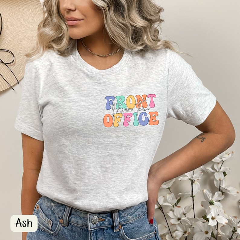 K&ouml;nnte beinhalten: Hellgraues T-Shirt mit dem Schriftzug "FRONT OFFICE" in bunten Buchstaben im Retro-Stil. Das Shirt hat einen Rundhalsausschnitt und kurze &Auml;rmel. Das Wort "Ash" ist unten links aufgedruckt.
